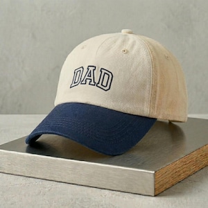May include: A beige baseball cap with a navy blue brim. The word "DAD" is embroidered in navy blue block letters on the front of the cap. The cap is sitting on a metallic surface.