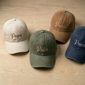 May include: Four baseball caps in cream, olive green, tan, and navy blue. Each cap features a curved brim, a button on the crown, and the word Papa embroidered in a cursive script on the front panel. The caps are arranged in a group.