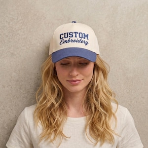 May include: A baseball cap featuring an off-white crown and a blue brim. The front of the cap displays the text CUSTOM Embroidery in dark blue thread, with the word CUSTOM in a bold block font and Embroidery in a cursive script font.