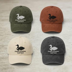 May include: Four baseball caps in olive green, rust orange, cream, and charcoal gray. Each cap features an embroidered graphic of a loon on water with the text "stupid canadian wolf bird" printed underneath in a simple, dark or light font.