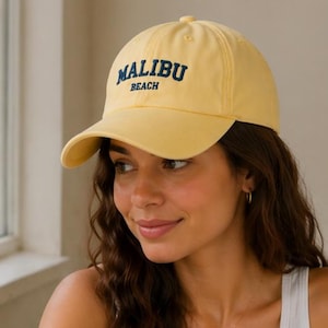 May include: A pale yellow baseball cap featuring the text MALIBU BEACH embroidered in dark blue capital letters on the front panel. The cap has a structured crown, a curved brim, and a button at the top center of the dome.