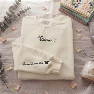 May include: A cream-colored crewneck sweatshirt featuring black embroidery. The chest displays the word Dream with four-pointed stars and a Mickey Mouse head icon. The sleeve features the text Dreams do come true with a small Mickey Mouse head icon.