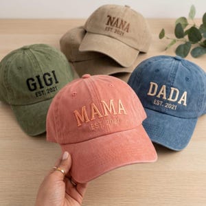 May include: A collection of baseball caps in various colors, including green, peach, blue, and tan. Each cap features embroidered text: "GIGI," "MAMA," "DADA," and "NANA," along with "EST. 2021." The caps are made of a soft, worn fabric.