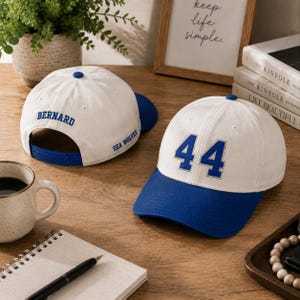May include: Two white baseball caps with blue brims and blue embroidery. One cap features the number 44 on the front. The other cap shows the name BERNARD on the back and the text SEA WOLVES on the side, with a blue adjustable snapback closure.