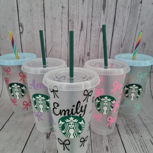 May include: Four clear plastic tumblers with lids and straws. Each tumbler features a black and white bow design and a green Starbucks logo. The tumblers are personalised with the names "Amy", "Emily", and two other names that are not visible in the image.