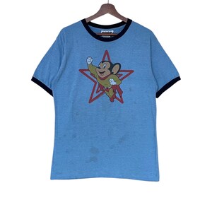 May include: Pale blue ringer t-shirt with a cartoon mouse superhero graphic inside a red star. The shirt has a black collar and sleeve trim. The graphic is in yellow, red, and green.