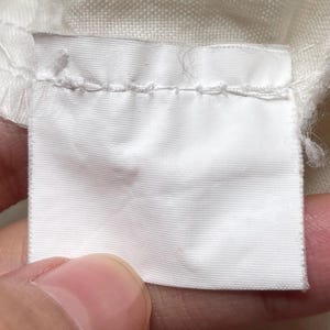 May include: Close-up of a white fabric label, rectangular in shape, with a blank surface. The label is attached to a piece of white fabric and is held by a person's fingers. The label has a stitched border.