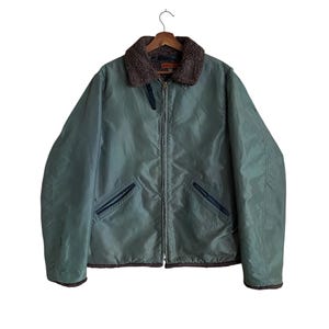 May include: A sage green jacket with a brown faux fur collar and trim, hanging on a wooden hanger. The jacket has a zipper closure, two front pockets, and a dark brown lining. The jacket is made of a smooth, possibly nylon, material.