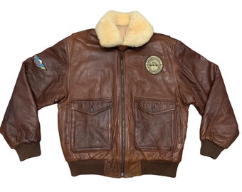 May include: A brown leather bomber jacket with a shearling collar and a zipper closure. The jacket has two pockets on the front and a patch on the left sleeve. The patch has a graphic of an airplane with the text "Wings of Freedom" on it.