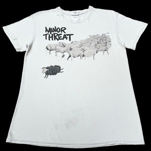 Op de afbeelding: Wit T-shirt met een zwart-witte afbeelding van een kudde schapen met &eacute;&eacute;n zwart schaap dat uit de kudde springt. De tekst "MINOR THREAT" is in zwart boven de schapen gedrukt.