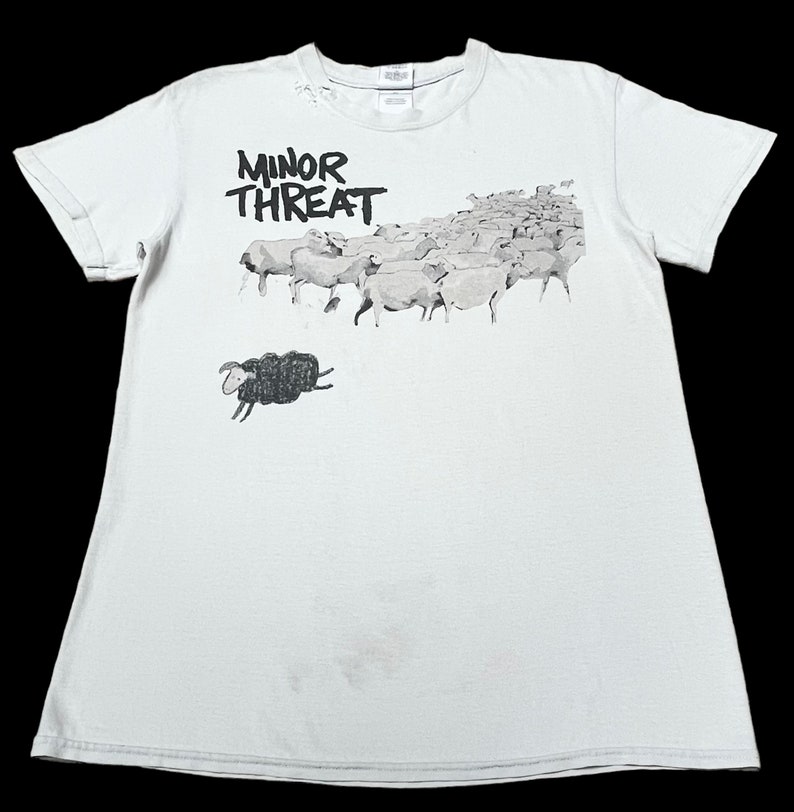 Op de afbeelding: Wit T-shirt met een zwart-witte afbeelding van een kudde schapen met &eacute;&eacute;n zwart schaap dat uit de kudde springt. De tekst "MINOR THREAT" is in zwart boven de schapen gedrukt.