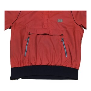 May include: A red pullover jacket with a navy blue elastic waistband and two zippered pockets. The jacket has a high neck and a small patch on the chest.