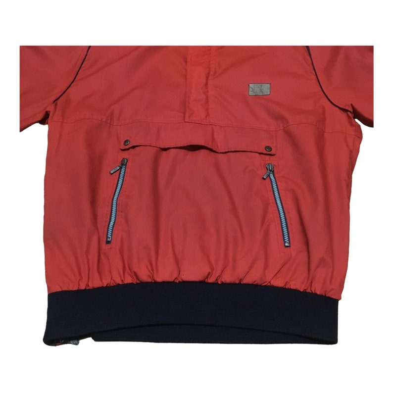 May include: A red pullover jacket with a navy blue elastic waistband and two zippered pockets. The jacket has a high neck and a small patch on the chest.