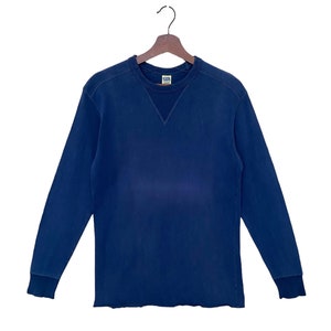 May include: A navy blue long-sleeved sweatshirt with a crew neck and a small, triangular detail at the neckline. The sweatshirt is hanging on a wooden hanger against a white background. The cuffs and hem are ribbed.