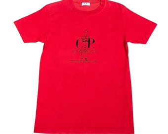 May include: Red short-sleeved t-shirt with a black graphic of a man standing with the text "C.P. COMPANY IDEAS FROM MASSIMO OSti"