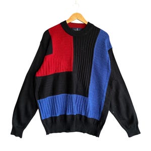 Può includere: Un maglione a blocchi di colore nero, rosso e blu. Il maglione presenta un design geometrico con sezioni a coste e a tinta unita. Le maniche e la parte inferiore del corpo sono nere a tinta unita. Il maglione è appeso a una gruccia di legno.