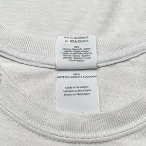 Op de afbeelding: Wit katoenen T-shirt label met wasinstructies en de tekst "100% COTTON/COTON/ALGODON", "Made in Nicaragua", "Fabriqu&eacute; au Nicaragua", "Hecho en Nicaragua".