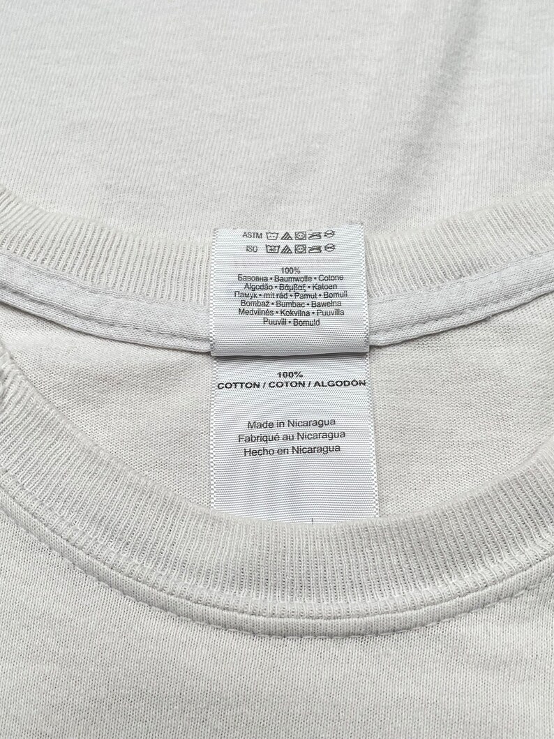 Op de afbeelding: Wit katoenen T-shirt label met wasinstructies en de tekst "100% COTTON/COTON/ALGODON", "Made in Nicaragua", "Fabriqu&eacute; au Nicaragua", "Hecho en Nicaragua".