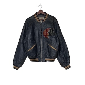 May include: A black leather bomber jacket with a brown and white striped collar and cuffs. The jacket has a zipper closure and a large embroidered patch on the left side with the letter "A" and a bird design.