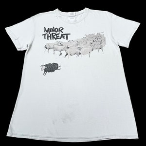 Op de afbeelding: Een lichtblauw T-shirt met een zwart-witte afbeelding van een zwart schaap dat wegrent van een kudde witte schapen. De tekst "MINOR THREAT" is in zwart op het shirt gedrukt.