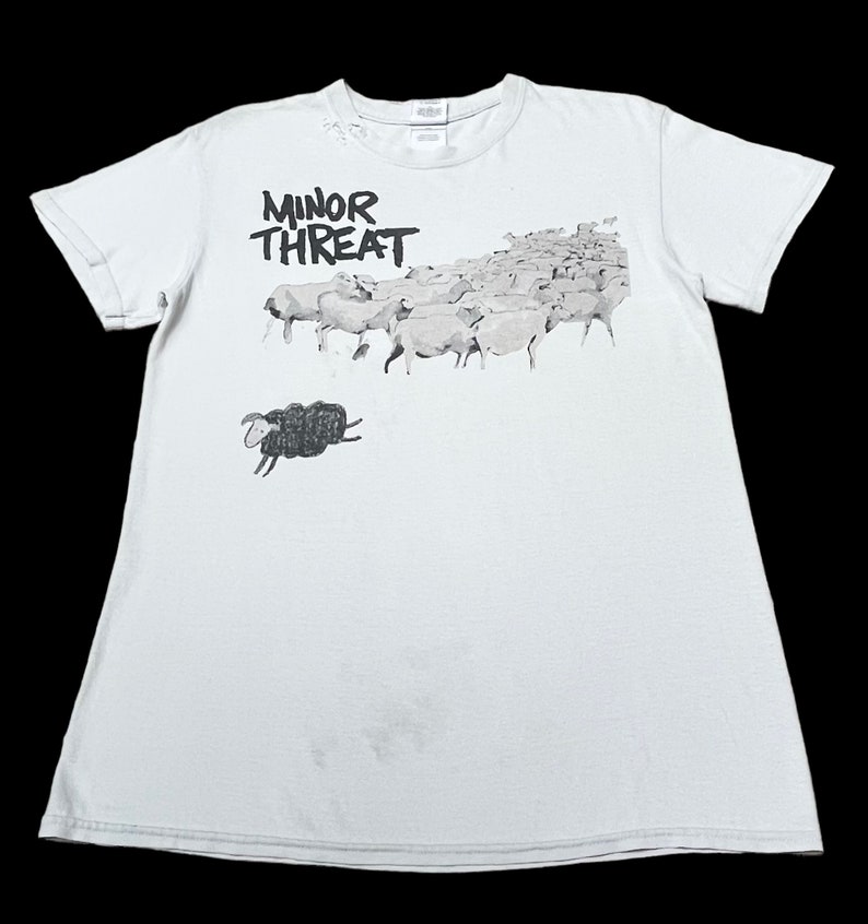 Op de afbeelding: Een lichtblauw T-shirt met een zwart-witte afbeelding van een zwart schaap dat wegrent van een kudde witte schapen. De tekst "MINOR THREAT" is in zwart op het shirt gedrukt.