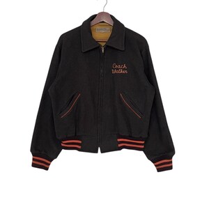 May include: A black wool jacket with a zip-up front, featuring orange trim on the pockets and cuffs. The jacket has the embroidered text "Coach Walker" in orange script on the chest. The jacket is hanging on a wooden hanger.