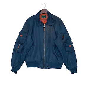 Puede incluir: Una chaqueta bomber azul oscuro con múltiples bolsillos y cierre de cremallera. La chaqueta tiene un forro naranja y una etiqueta verde con el texto "Flight Gear" en el interior.