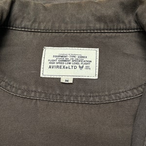 Puede incluir: Una chaqueta vaquera negra con una etiqueta blanca que dice "Certificate of Authenticity", "EQUIPMENT-TYPE-AVIREX", "STOCK NO. 28-A6755", "FLIGHT GARMENT SPECIFICATION", "HIGH SPEED LOW LEVEL FLIGHT", "AVIREX LTD", "EST 1975" y "M".