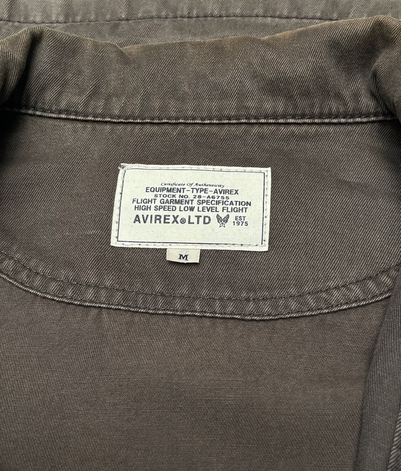 Puede incluir: Una chaqueta vaquera negra con una etiqueta blanca que dice "Certificate of Authenticity", "EQUIPMENT-TYPE-AVIREX", "STOCK NO. 28-A6755", "FLIGHT GARMENT SPECIFICATION", "HIGH SPEED LOW LEVEL FLIGHT", "AVIREX LTD", "EST 1975" y "M".