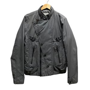 May include: A charcoal gray jacket with a unique asymmetrical design. The jacket features a stand-up collar, multiple pockets with zippers, and decorative buckles. It is hanging on a wooden hanger against a white background.