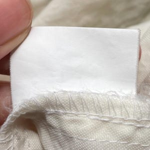 May include: Close-up of a white rectangular fabric label, held by a person's fingers, attached to a beige fabric. The label is blank, with a smooth, clean surface. The beige fabric has visible stitching.