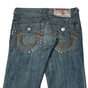 May include: A pair of blue denim jeans with a faded wash and distressed details. The back pockets feature a signature embroidered horseshoe design with colorful stitching. The jeans have a "True Religion" label with the text "World Tour" on the back.