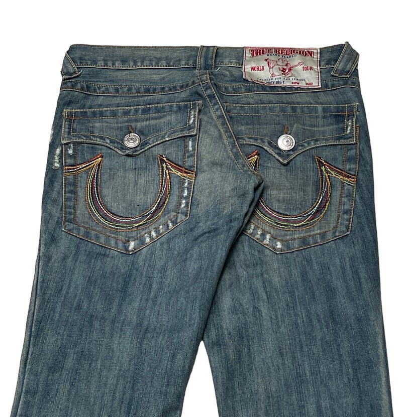 May include: A pair of blue denim jeans with a faded wash and distressed details. The back pockets feature a signature embroidered horseshoe design with colorful stitching. The jeans have a "True Religion" label with the text "World Tour" on the back.