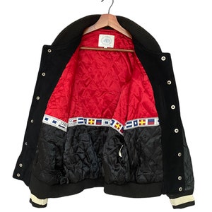 May include: Black varsity jacket with a red quilted lining and a white and blue striped trim. The jacket has a label that says "J. Press" on the inside.