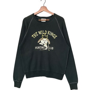 May include: A dark green sweatshirt featuring a yellow graphic of a dog wearing a crown and holding a bow and arrow. The text "The Wild Kings Hunting Club" is printed below the dog.