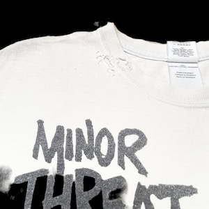Op de afbeelding: Wit T-shirt met een verweerde look, met de tekst "MINOR THREAT" in zwarte letters.