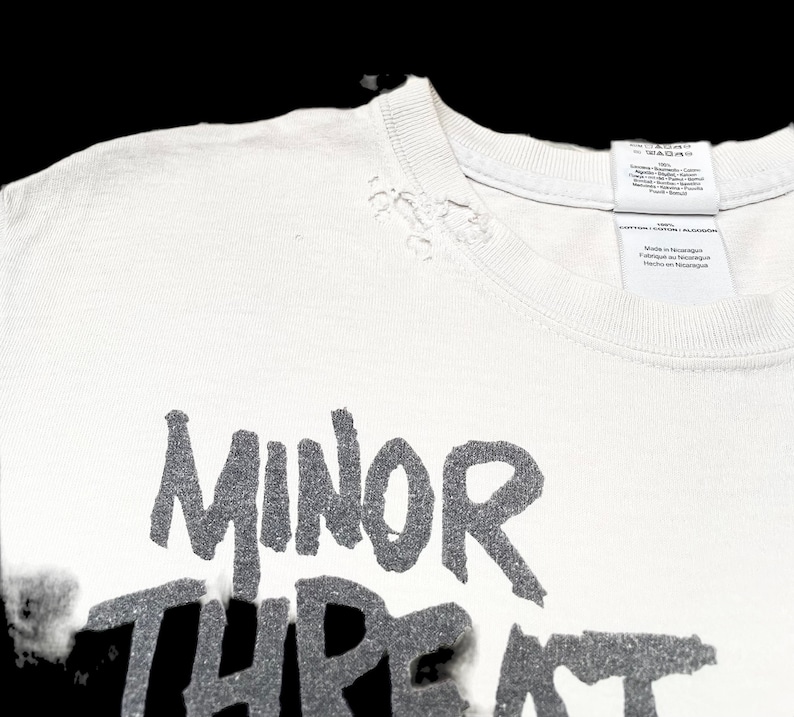 Op de afbeelding: Wit T-shirt met een verweerde look, met de tekst "MINOR THREAT" in zwarte letters.