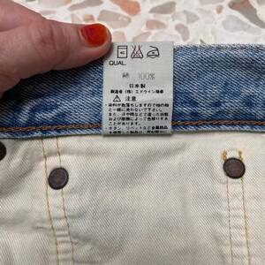 以下が含まれることがあります： 青いデニム生地のラベルのクローズアップ。白いテキストとケアの説明が書かれています。ラベルには「QUAL. 100% 日本製 (株) エドウイン商事  ▲注意 ・染料が色落ちしますので他の物と一緒に洗わないで下さい。 また、汗や顔などで湿った状態および摩擦によって色移りする事があります。 ・ボタン、リベットなど金属部品を使用した製品は、突起部分に」と記載されています。