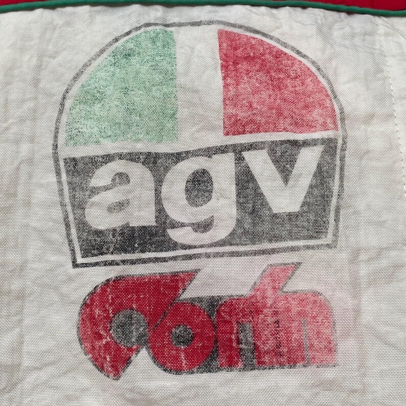 May include: Close-up of a white fabric with a faded AGV logo. The logo features the Italian flag colors, green, white, and red, above the letters "AGV" in white. Below the letters is the word "com" in red.