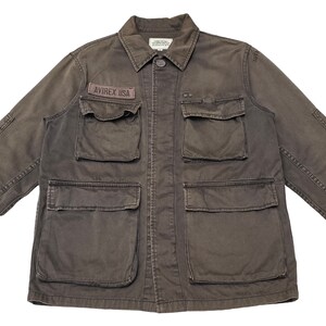 Puede incluir: Una chaqueta utilitaria marr&oacute;n oscuro con m&uacute;ltiples bolsillos y un parche que dice "AVIREX USA".