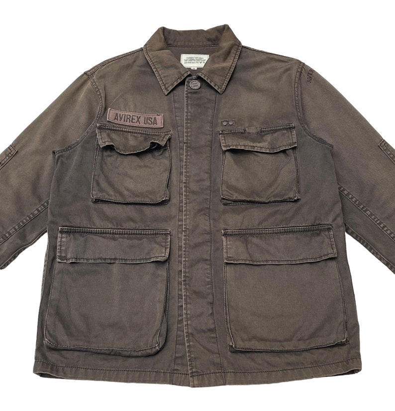 Puede incluir: Una chaqueta utilitaria marr&oacute;n oscuro con m&uacute;ltiples bolsillos y un parche que dice "AVIREX USA".