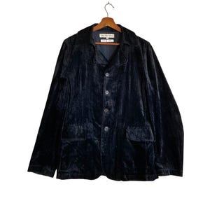 Pode incluir: Um blazer de veludo preto com gola entalhada, fecho de quatro botões e dois bolsos frontais. O casaco está pendurado numa cabide de madeira sobre um fundo branco. O blazer tem um design clássico e sob medida.