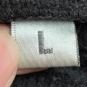 May include: A white tag with a black "L" printed on it. The tag is attached to a black garment.