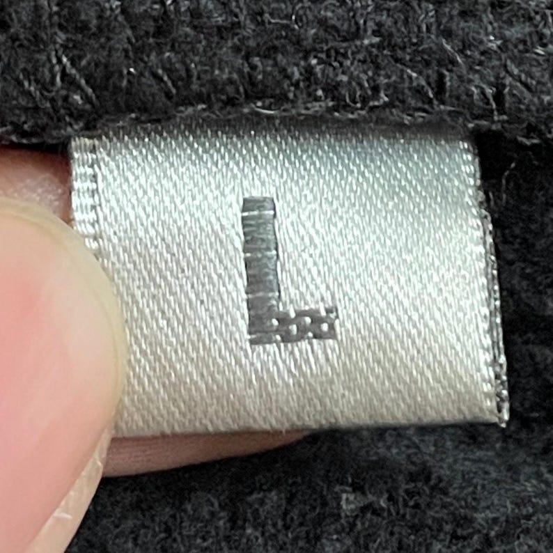 May include: A white tag with a black "L" printed on it. The tag is attached to a black garment.