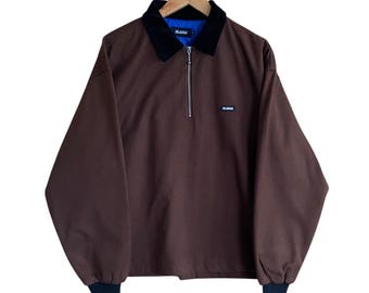 May include: Brown long-sleeve jacket with a black collar and a silver zipper. The jacket has a small logo on the chest and is hanging on a wooden hanger. The cuffs are black. The brand name "XLARGE" is visible on the jacket.