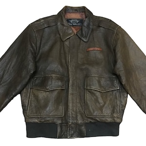 May include: A brown leather bomber jacket with a zipper closure and two flap pockets. The jacket has a ribbed knit collar, cuffs, and waistband. The jacket has a patch on the left chest that says "The Buckle".