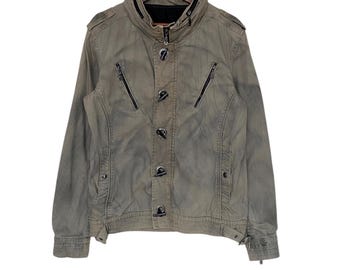 May include: A faded olive green jacket with a stand-up collar and black zippers. The jacket features a button-up front with unique toggle closures and a tailored fit. The jacket is hanging on a wooden hanger.