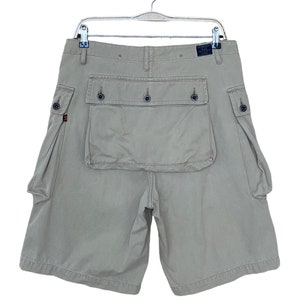 May include: Beige cargo shorts with multiple pockets, including a large flap pocket on the back. The shorts have a button fly and belt loops.