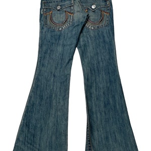 May include: A pair of blue denim jeans with a wide leg opening. The jeans have a rainbow-colored stitching detail on the back pockets and a True Religion brand label.