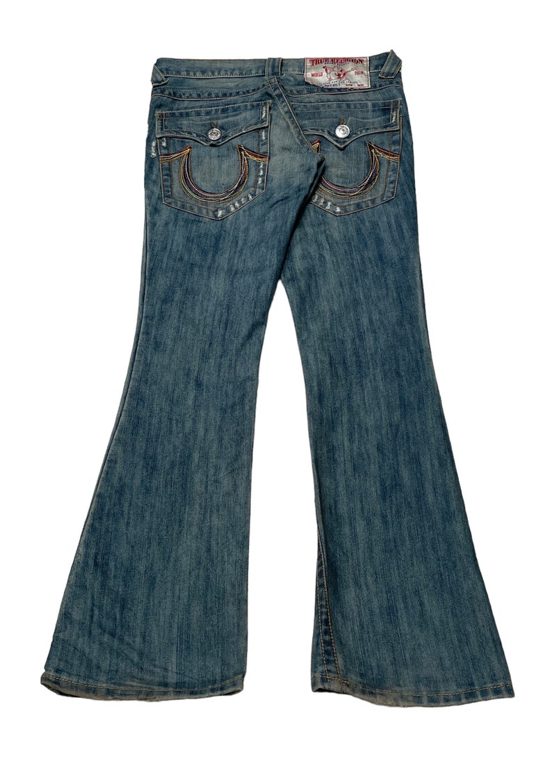May include: A pair of blue denim jeans with a wide leg opening. The jeans have a rainbow-colored stitching detail on the back pockets and a True Religion brand label.